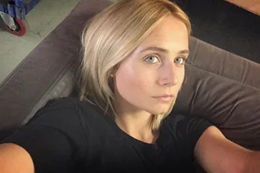 A person with blonde hair takes a selfie lying on a couch, wearing a black shirt and looking at the camera.