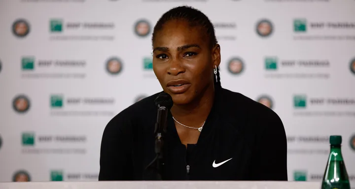 Tennis player speaking at a press conference, wearing a black Nike jacket, with logos in the background.