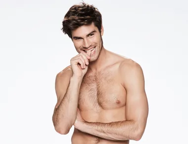 Man with tousled hair, shirtless, smiling playfully while touching his lips with his hand against a plain background.