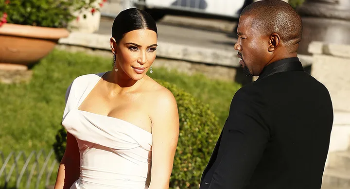 Two people in formal attire outdoors, one in a white dress, the other in a black suit, conversing on a sunny day.