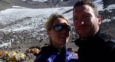 Couple takes a selfie in front of snowy mountain landscape with tents in the background.