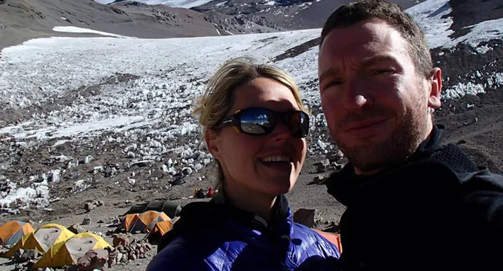 Couple takes a selfie in front of snowy mountain landscape with tents in the background.