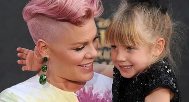A woman with pink hair smiles while holding a young girl in a black dress, both looking at each other fondly.