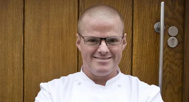 Chef in white coat and glasses smiling in front of a wooden door.