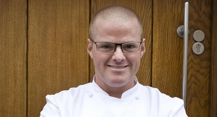 Chef in white coat and glasses smiling in front of a wooden door.