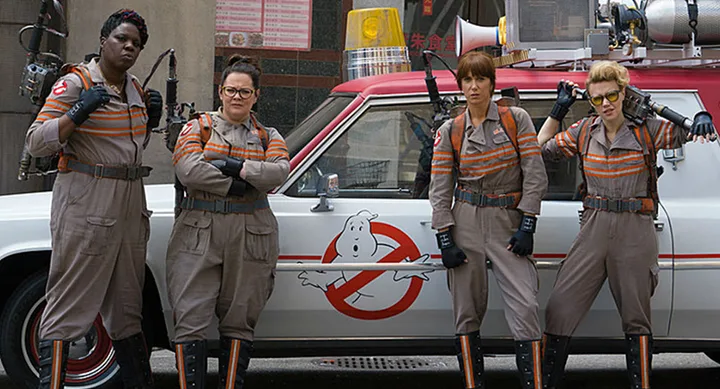 Four women in Ghostbusters suits stand confidently in front of the Ecto-1 vehicle from the 2016 "Ghostbusters" film.