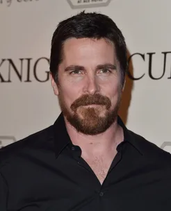 A person with a beard in a black shirt standing in front of a wall with partially visible text.