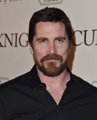 A person with a beard in a black shirt standing in front of a wall with partially visible text.