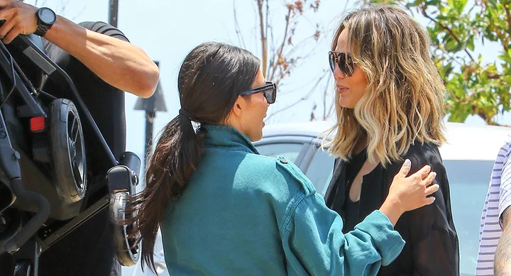 Two women in sunglasses talking and smiling outside, with one reaching out to touch the other's shoulder.