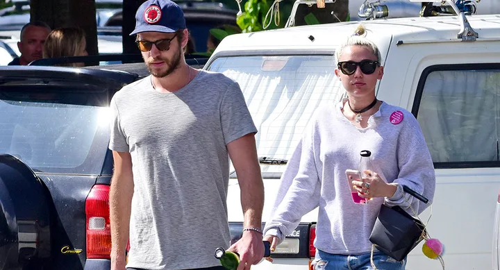 Two people walking outdoors. The man wears a cap and gray shirt; the woman has sunglasses, a light sweater, and holds a drink.