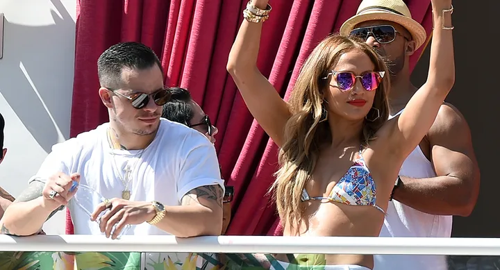 A woman in a bikini and sunglasses raises her hands at a pool party, surrounded by people in casual summer attire.