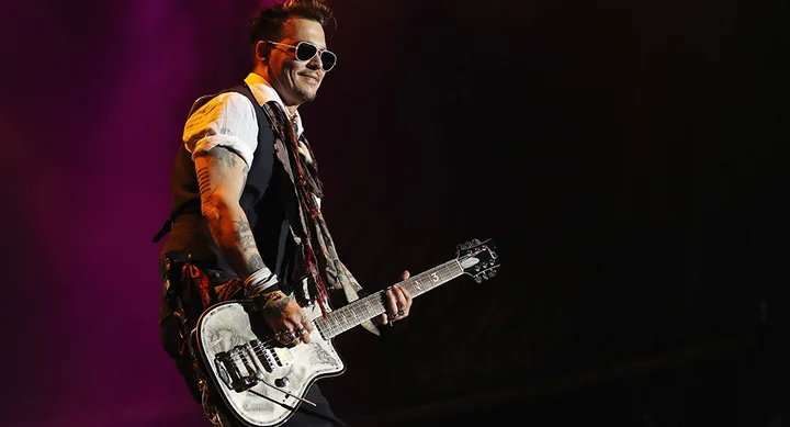 Musician playing guitar on stage, wearing sunglasses, vest, and scarf, with a dark background.