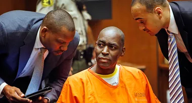 Man in orange jail uniform sits, flanked by two men in suits, in a courtroom setting.