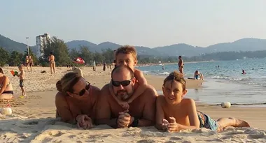 Family of four lying on the sandy beach, ocean and mountains in the background.
