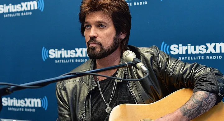 Musician playing guitar during SiriusXM interview, with microphone and studio backdrop visible.