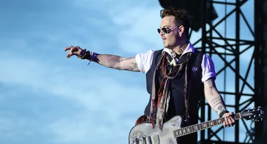 Musician wearing sunglasses, playing guitar on stage with blue sky background.