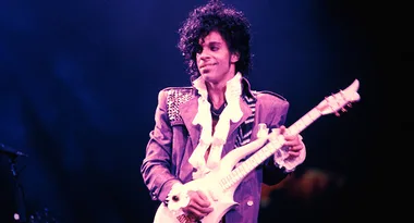 Musician in a purple jacket plays an electric guitar on stage.