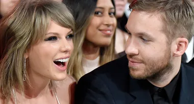 Two people sitting closely, smiling and talking at an event, with blurred audience in the background.