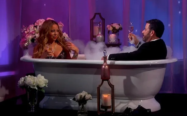 Two people in a bubble-filled bathtub, toasting with champagne in a room decorated with flowers and candles.