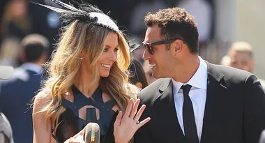 A couple in formal attire smiling at each other, with the woman wearing a feathered fascinator.