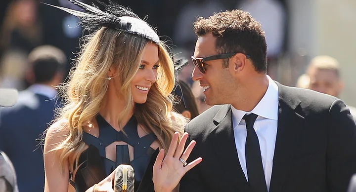 A couple in formal attire smiling at each other, with the woman wearing a feathered fascinator.