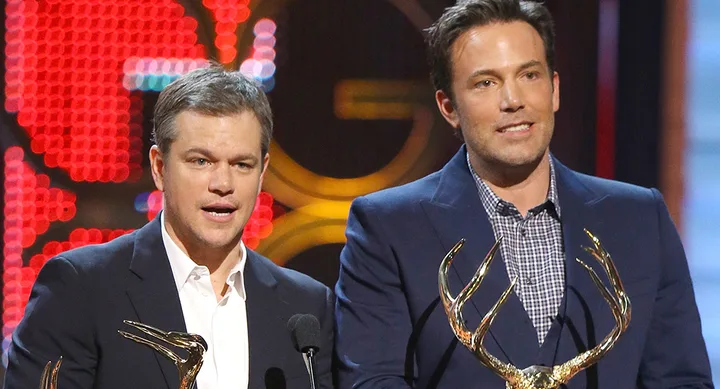 Two men in suits holding gold awards, speaking at a podium during an event.