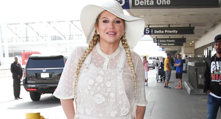 Woman in a white embroidered outfit and hat with braids, standing at an airport terminal.