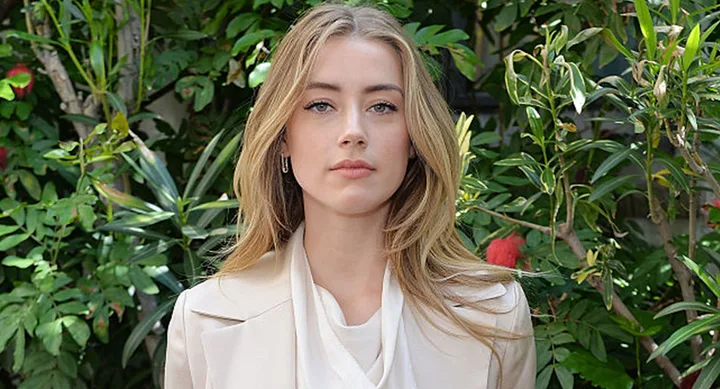 A person with long blonde hair stands in front of green foliage, wearing a light-colored jacket and white top.