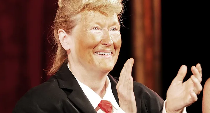A person in a suit and tie with exaggerated orange facial makeup and fake hair clapping.