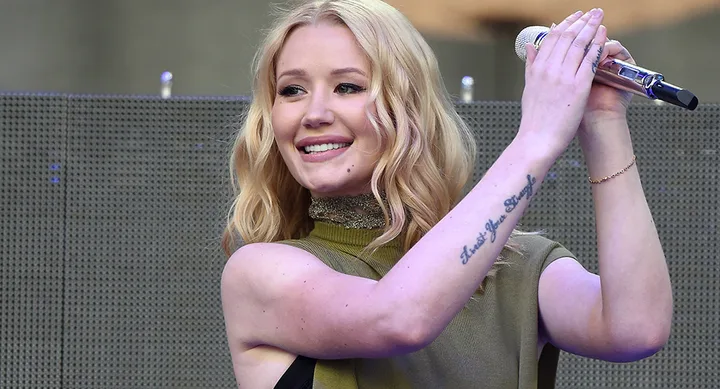 Singer with blonde hair holding a microphone on stage, smiling, with a tattoo visible on her arm.