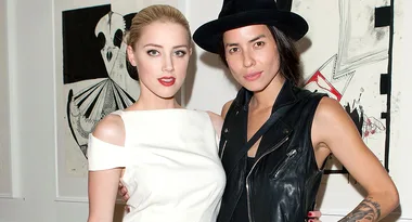 Two people posing together in front of abstract artwork, one in a white dress, the other in a black hat and vest.