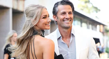 Couple smiling outdoors, woman in black dress, man in white suit jacket.