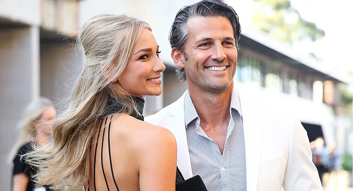Couple smiling outdoors, woman in black dress, man in white suit jacket.