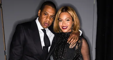 Two individuals posing together, one in a black suit, the other in a black dress, smiling at the camera.