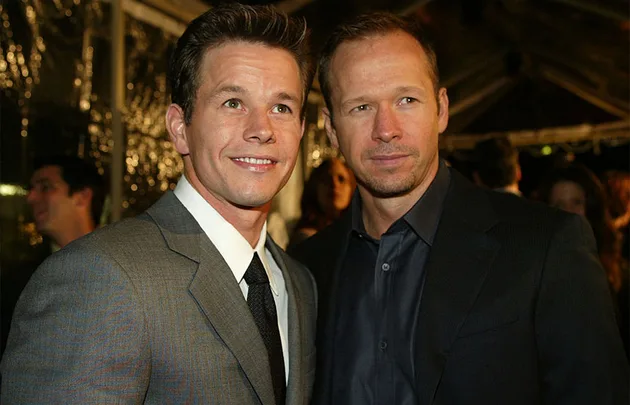 Two men in suits pose together at an event, with dim lighting and people in the background.