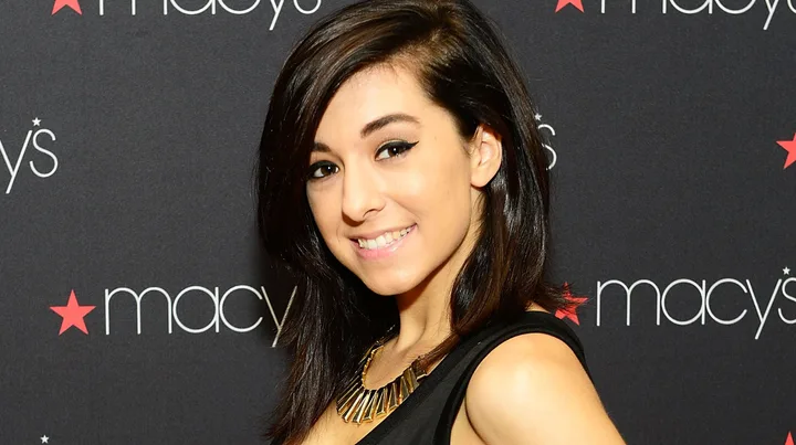 A person smiles at a Macy's event, wearing a black dress and a gold necklace, with long dark hair.
