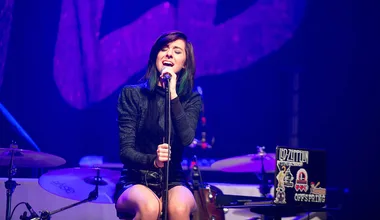 Singer performing on stage with microphone, seated next to drums and decorated laptop.