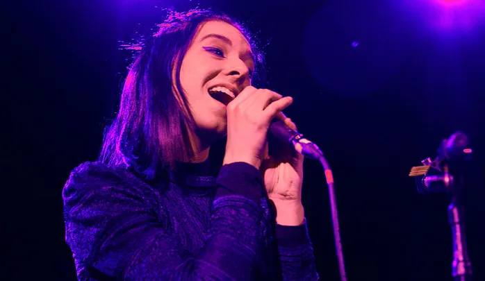 Singer performing on stage under purple lighting, holding a microphone, with intense expression.