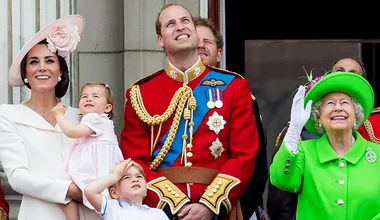 A family dressed in formal attire stands on a balcony, smiling and looking up at the sky.