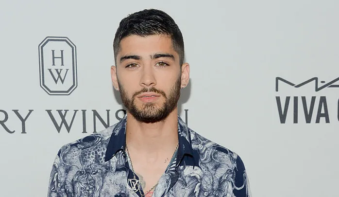 Young man with short dark hair and a beard, wearing a patterned shirt, standing in front of a step and repeat backdrop.