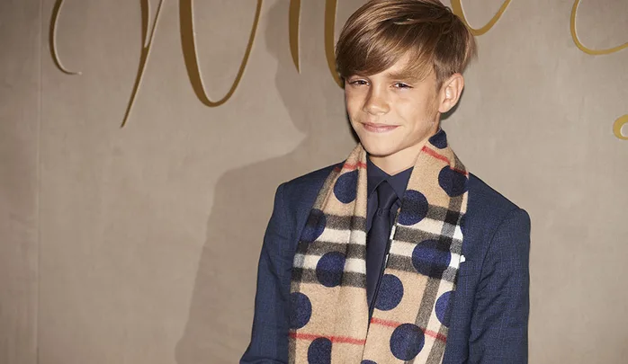 A young boy in a blue suit and tie, wearing a beige plaid scarf with blue polka dots, stands smiling against a beige wall.