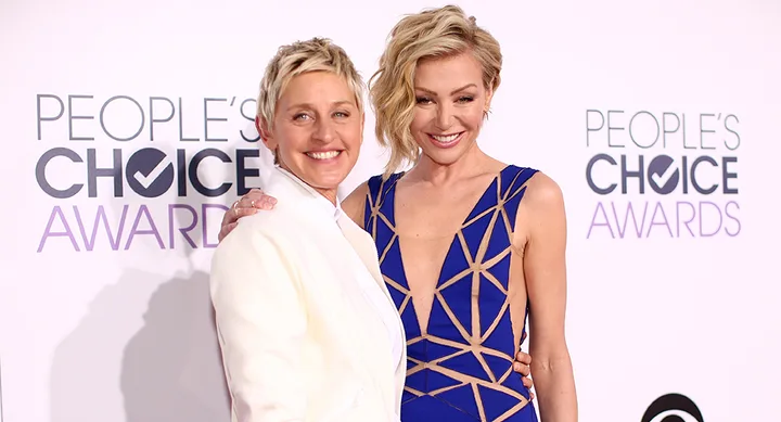 Two people smiling on the red carpet at the People's Choice Awards; one in a white suit, the other in a blue patterned dress.