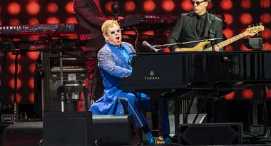 Musician in a blue suit plays a Yamaha piano on stage with a band, red lights in the background.
