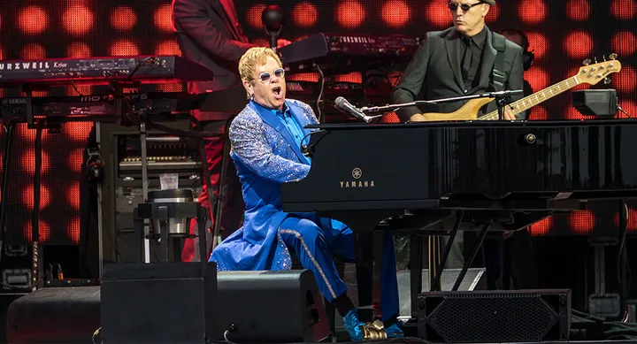 Musician in a blue suit plays a Yamaha piano on stage with a band, red lights in the background.