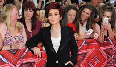 Sharon Osbourne poses with fans at an outdoor event, some holding phones, on a red X Factor-themed backdrop.