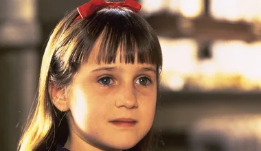 Young girl with brown hair in a red bow, looking thoughtful against a blurred background.