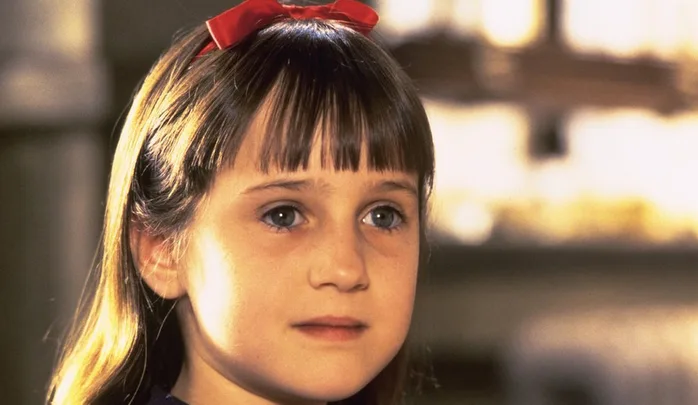 Young girl with brown hair in a red bow, looking thoughtful against a blurred background.