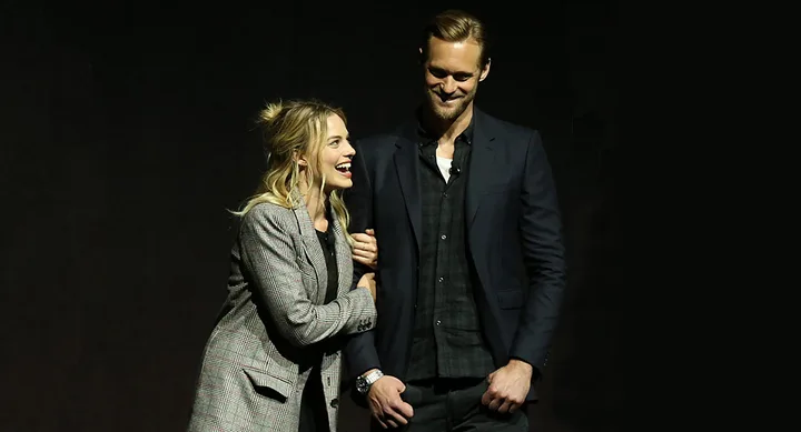 Two people smiling on stage, woman in plaid blazer links arm with man in dark suit.