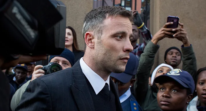 A person in a suit is surrounded by media and onlookers, some holding phones, outside a building.