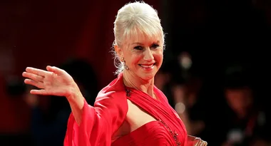 A person with white hair in a red dress waves with a smile, standing on a stage or red carpet with blurred background.